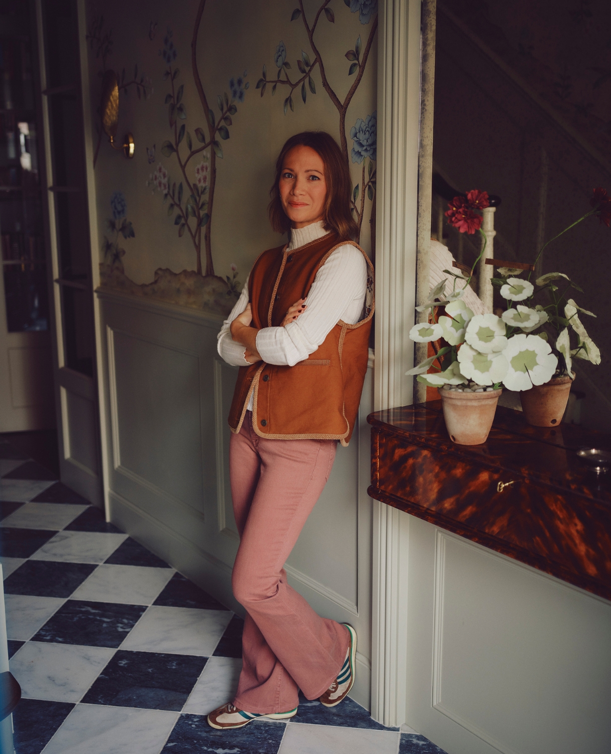 A woman stands indoors against a wall with floral wallpaper, wearing a brown vest, white sweater, and pink pants. She is smiling with arms crossed. A vase with white flowers sits on a table beside her. The floor is black and white checkered.
