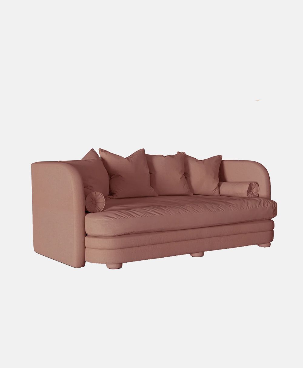 A modern, plush pink sofa with rounded arms, four square back cushions, and two cylindrical side pillows, set against a plain white background.