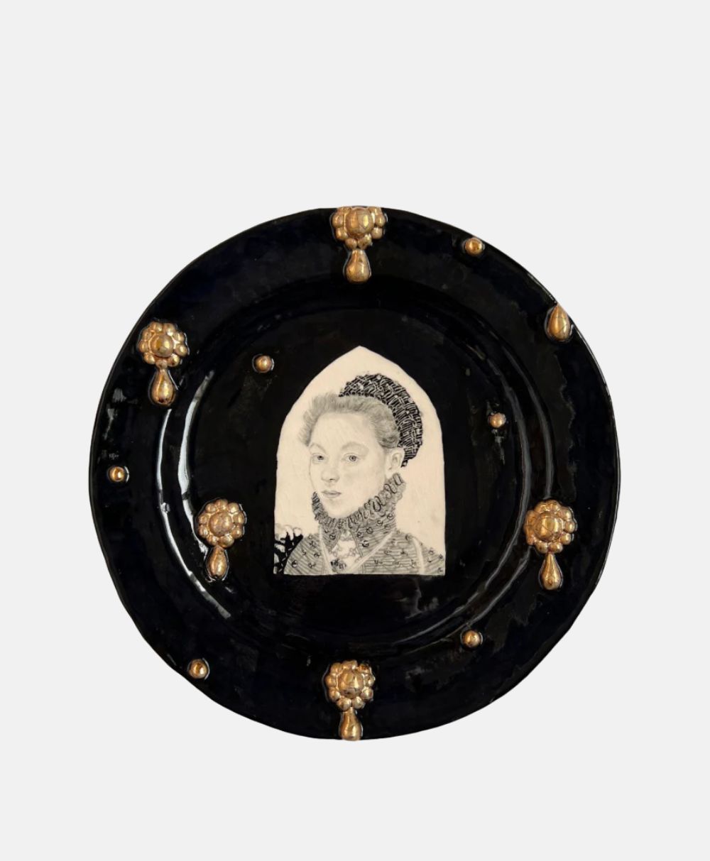 A black ceramic plate decorated with gold accents features a monochrome portrait of a woman in historical attire at its center.
