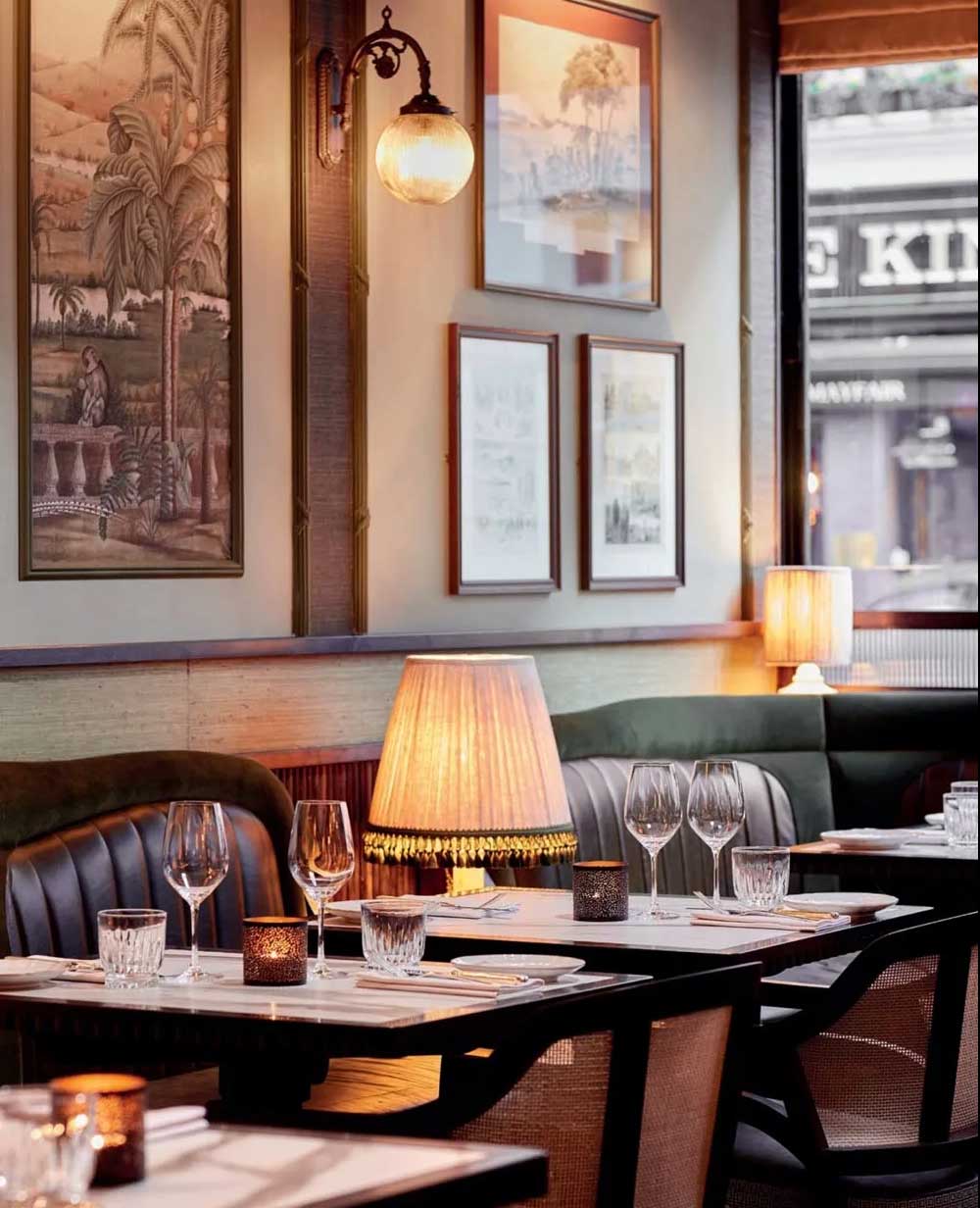 Elegant restaurant interior with softly lit tables, set with wine glasses and candles. Wall art and framed pictures decorate the background, and a window shows a blurred street view outside. Cozy and inviting atmosphere.