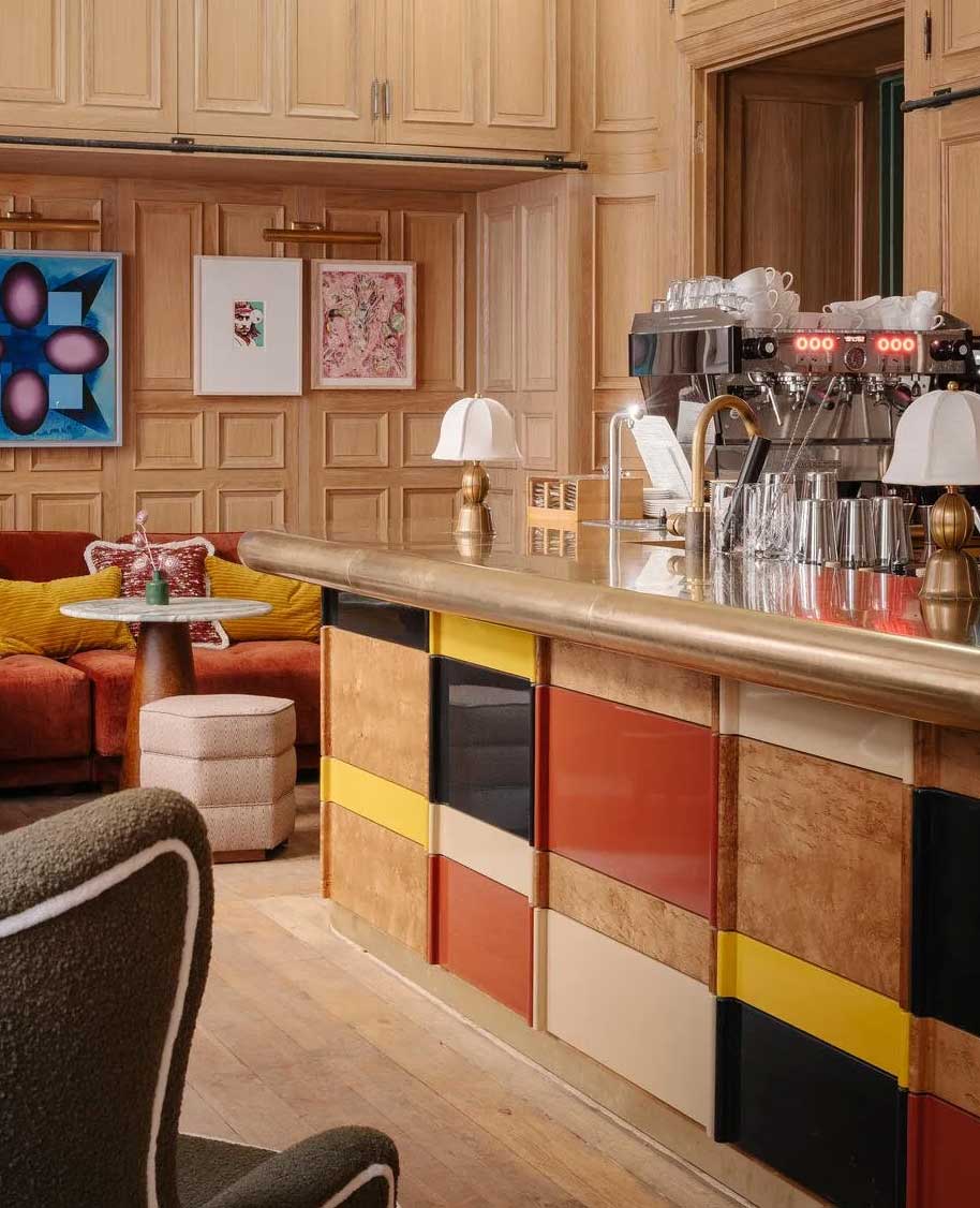 A cozy cafe with a colorful counter, coffee machines, a small round table with chairs, wall art, wood-paneled walls, and warm lighting.
