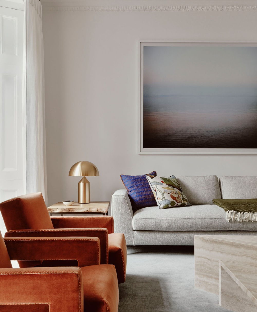 Modern living room with a light gray sofa, colorful cushions, two rust-colored armchairs, a marble coffee table, a gold table lamp, and a large abstract artwork on the wall. Sunlight streams through a window with sheer curtains.
