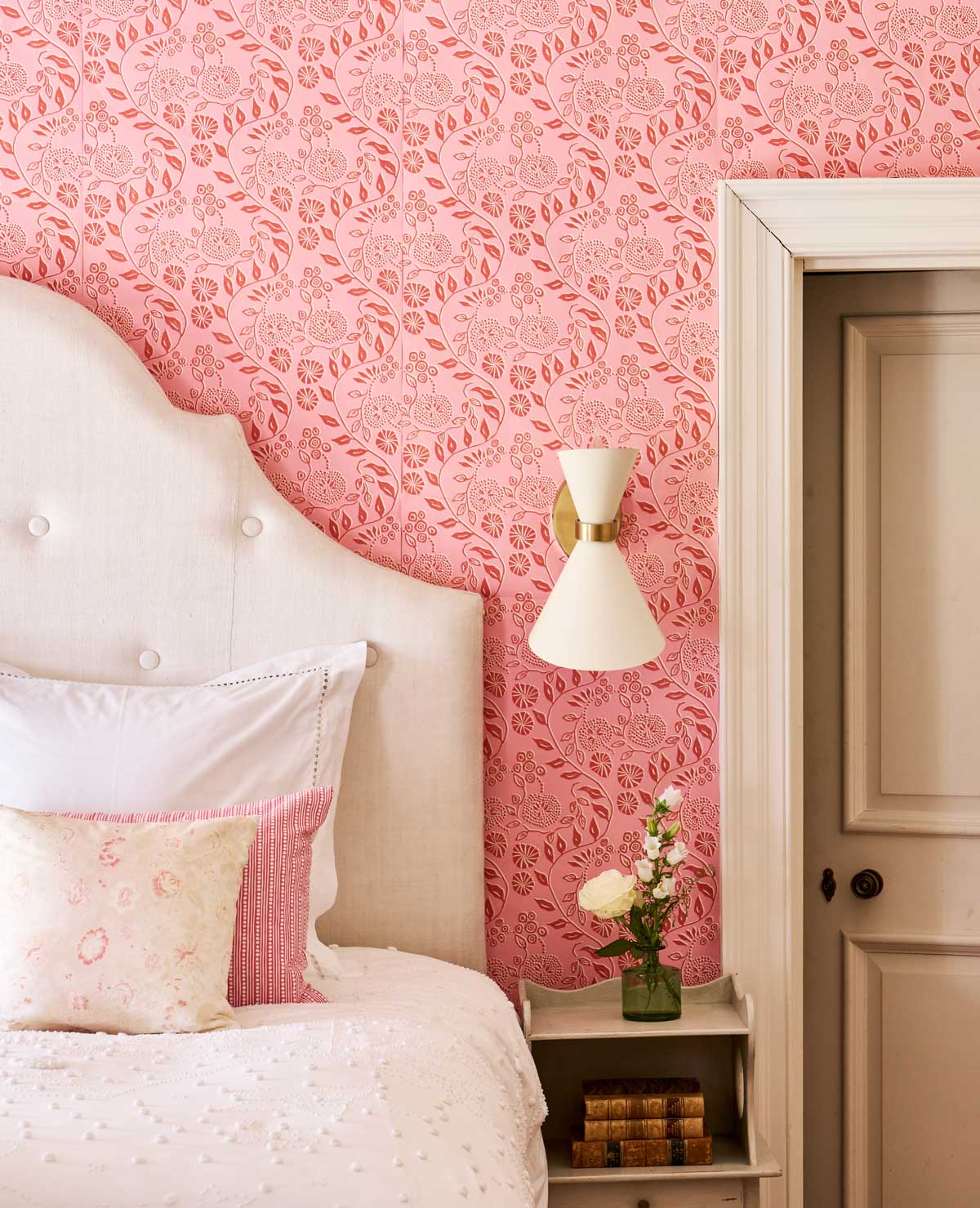 Bloomfield Ink wallpaper in an Elegant bedroom corner with a tufted beige headboard, pink floral wallpaper, white bedding with pink accents, a nightstand holding flowers, books, and a modern wall sconce above.