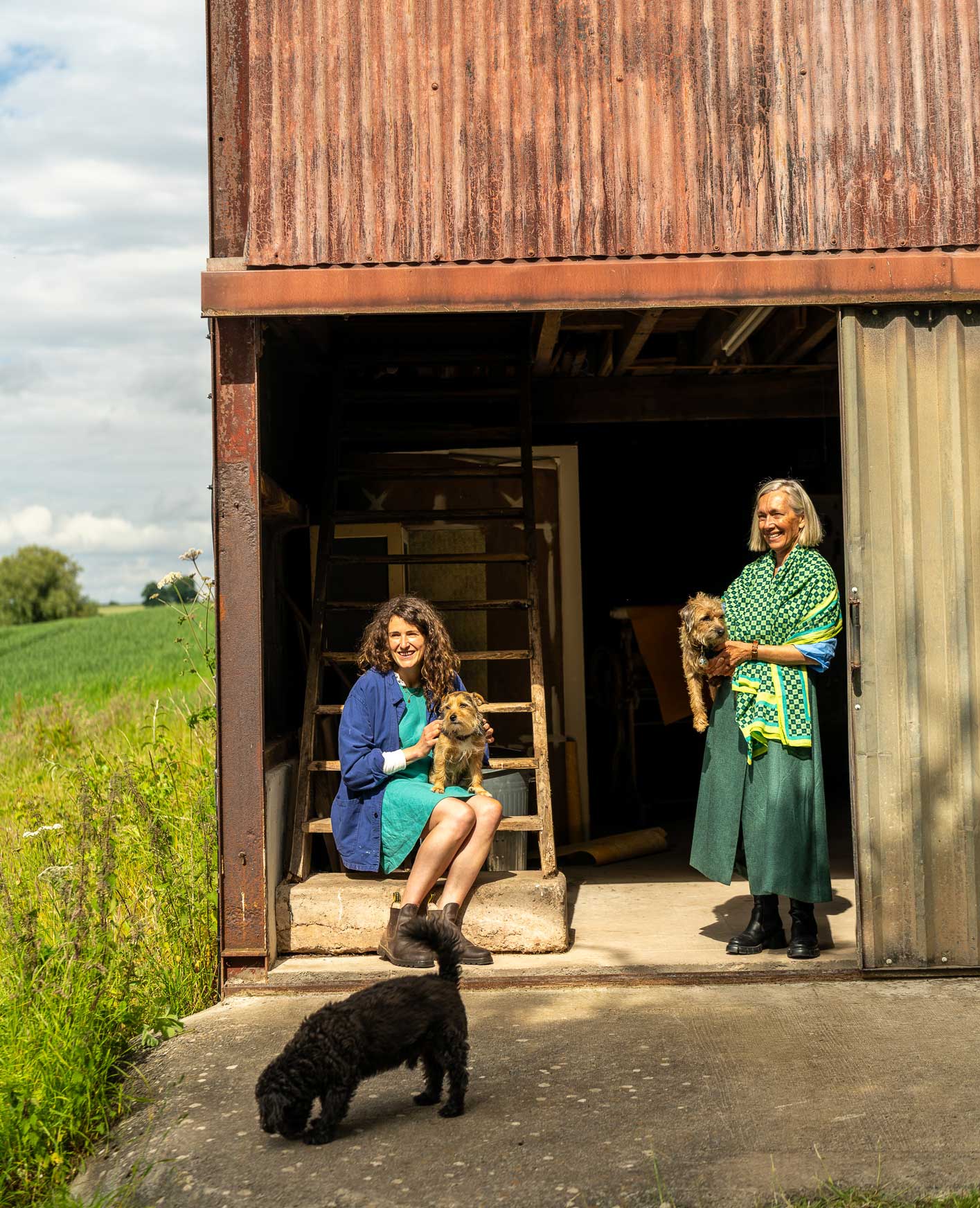 Bloomfielf Ink founders stand and sit in front of a barn, each holding a small dog. Another dog walks in the foreground. Green fields and blue sky are visible in the background.