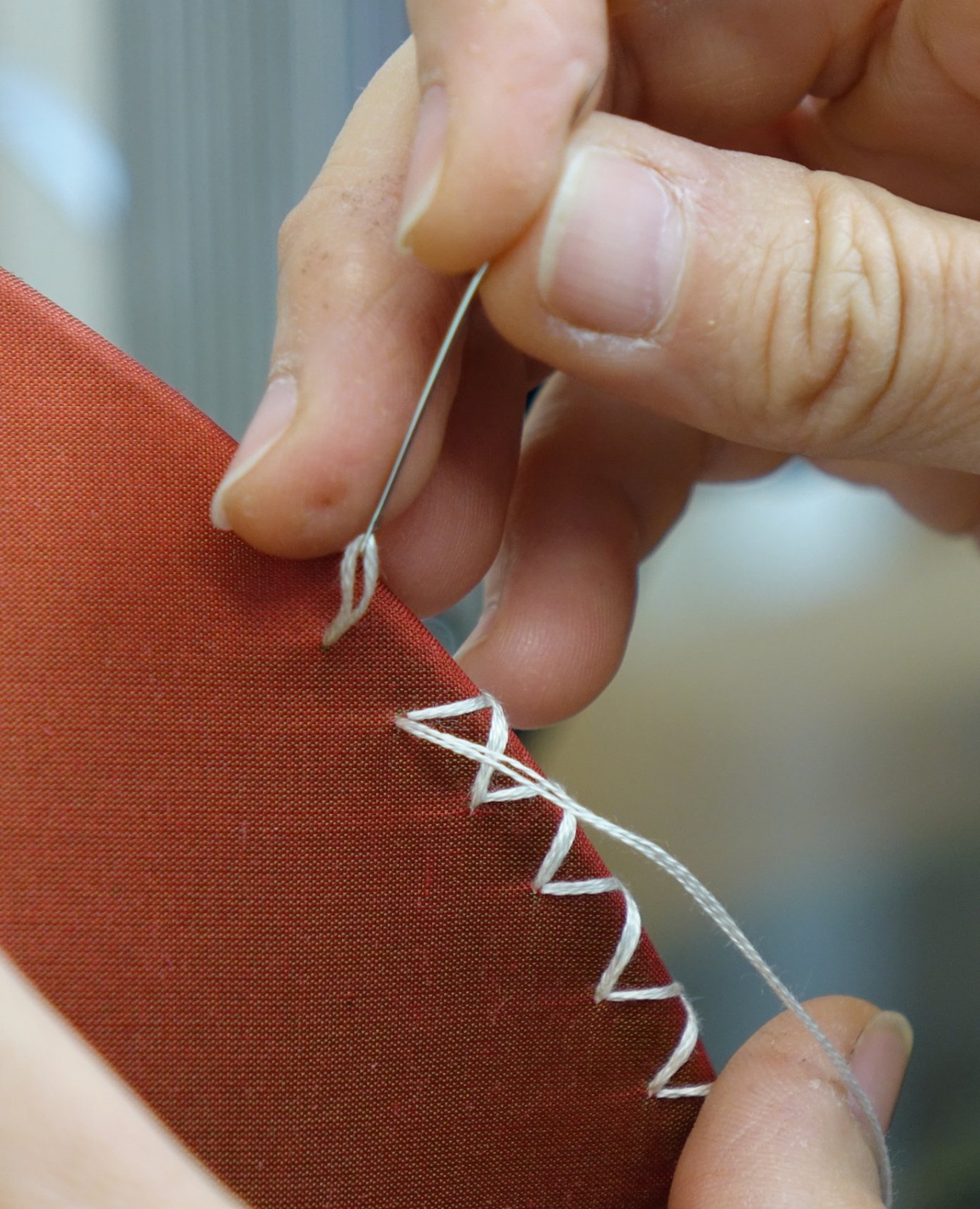 A craftsperson hand-stitching an ivory zigzag border along the edge of a terracotta fabric lampshade by Fosbery Studio, Scheme UK