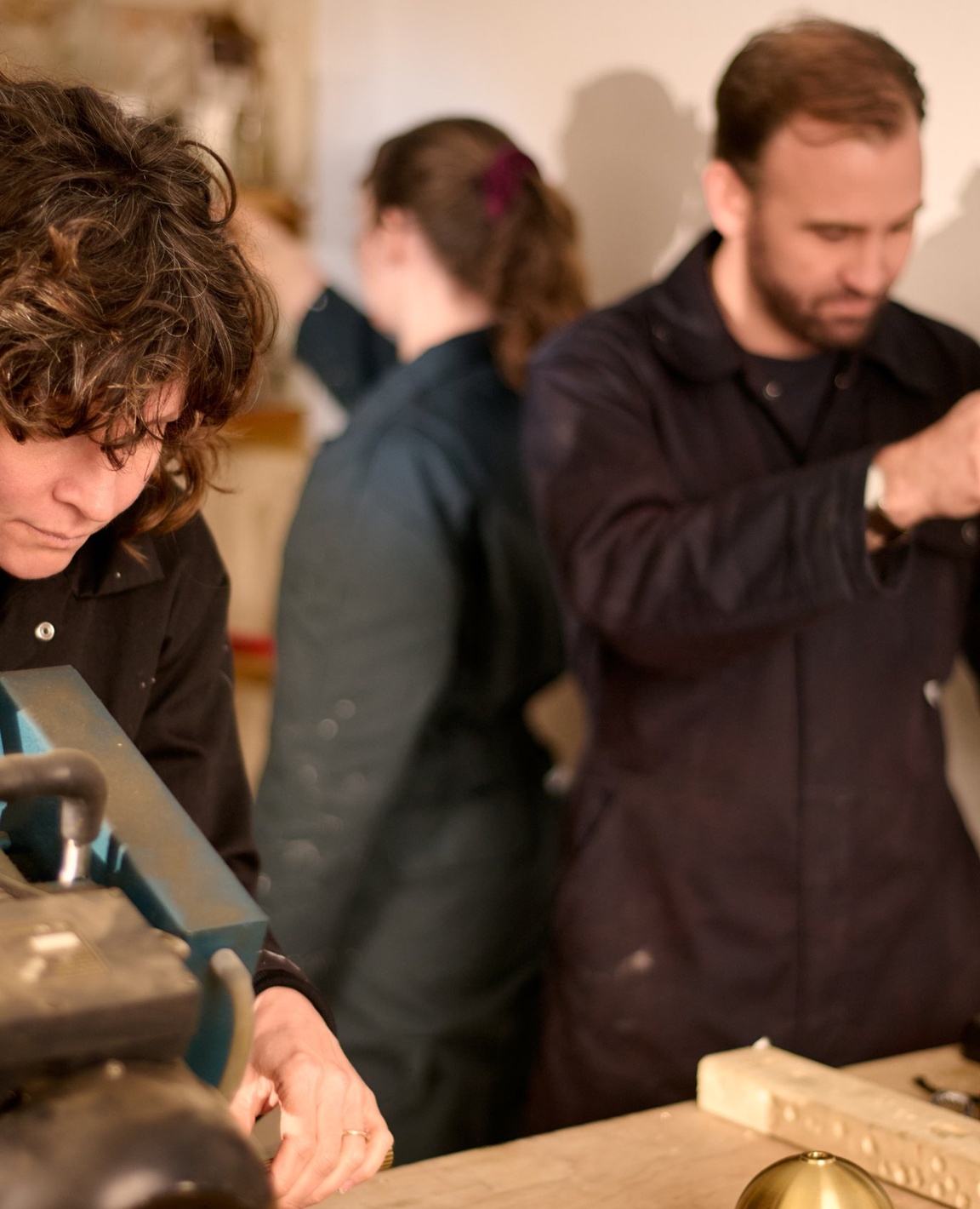 Makers in dark overalls at their benches in the Fosebery workshop, one working at a machine in the foreground and another at a drill behind. Fosbery Studio, Scheme UK