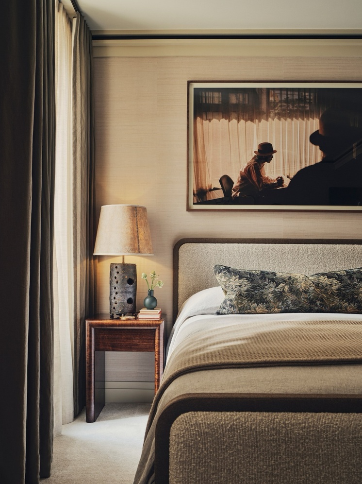 A bedroom corner designed by Christian Bense, with a boucle-upholstered bed in warm taupe, a floral-print cushion, a wood and ceramic table lamp on a dark wood nightstand, and a framed photograph hung above the headboard against beige grasscloth wallpaper.