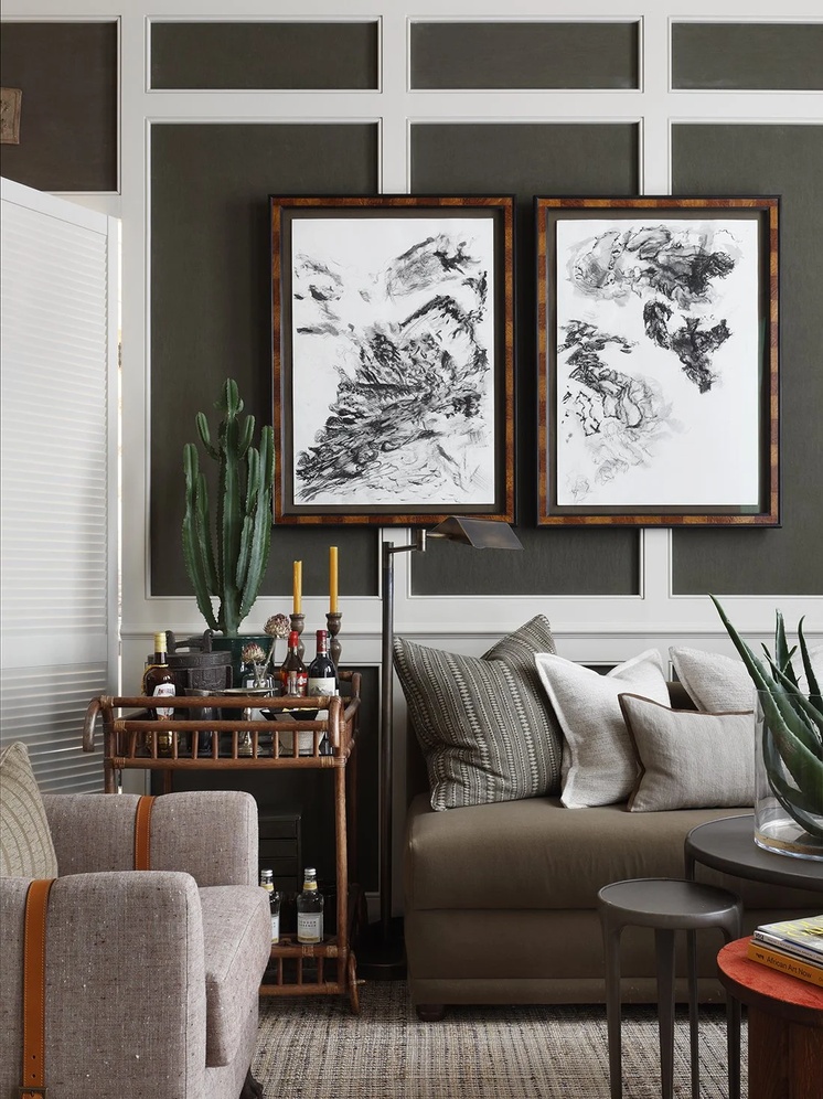 A snug corner by interior designer Christian Bense, with olive green wall panelling, two framed charcoal artworks above a low sofa with striped and plain cushions, a rattan drinks trolley, a floor lamp, and a leather-trimmed armchair in the foreground.