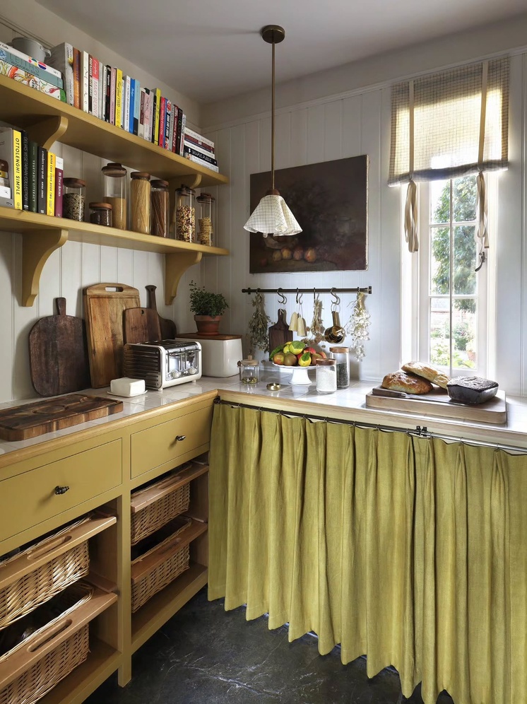 A country-style pantry designed by Christian Bense, with mustard-yellow cabinetry and open shelving stacked with cookbooks and storage jars, a woven basket drawer unit, a gingham roman blind at the window, and a sage-green curtain skirting the counter below.