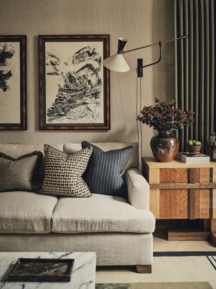 A living room by Christian Bense, with a beige linen sofa styled with patterned and striped cushions beneath a large charcoal abstract drawing in a tortoiseshell frame, next to an oak side table with a studio pottery vase and a swing-arm wall light.